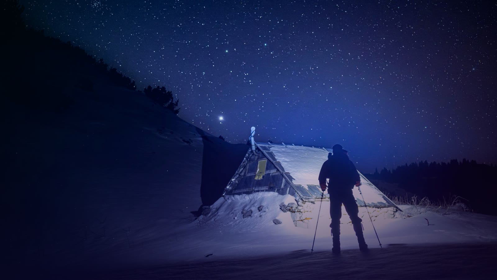 Silhouette of a skier standing by a snow-covered tent under a starry night sky in winter.