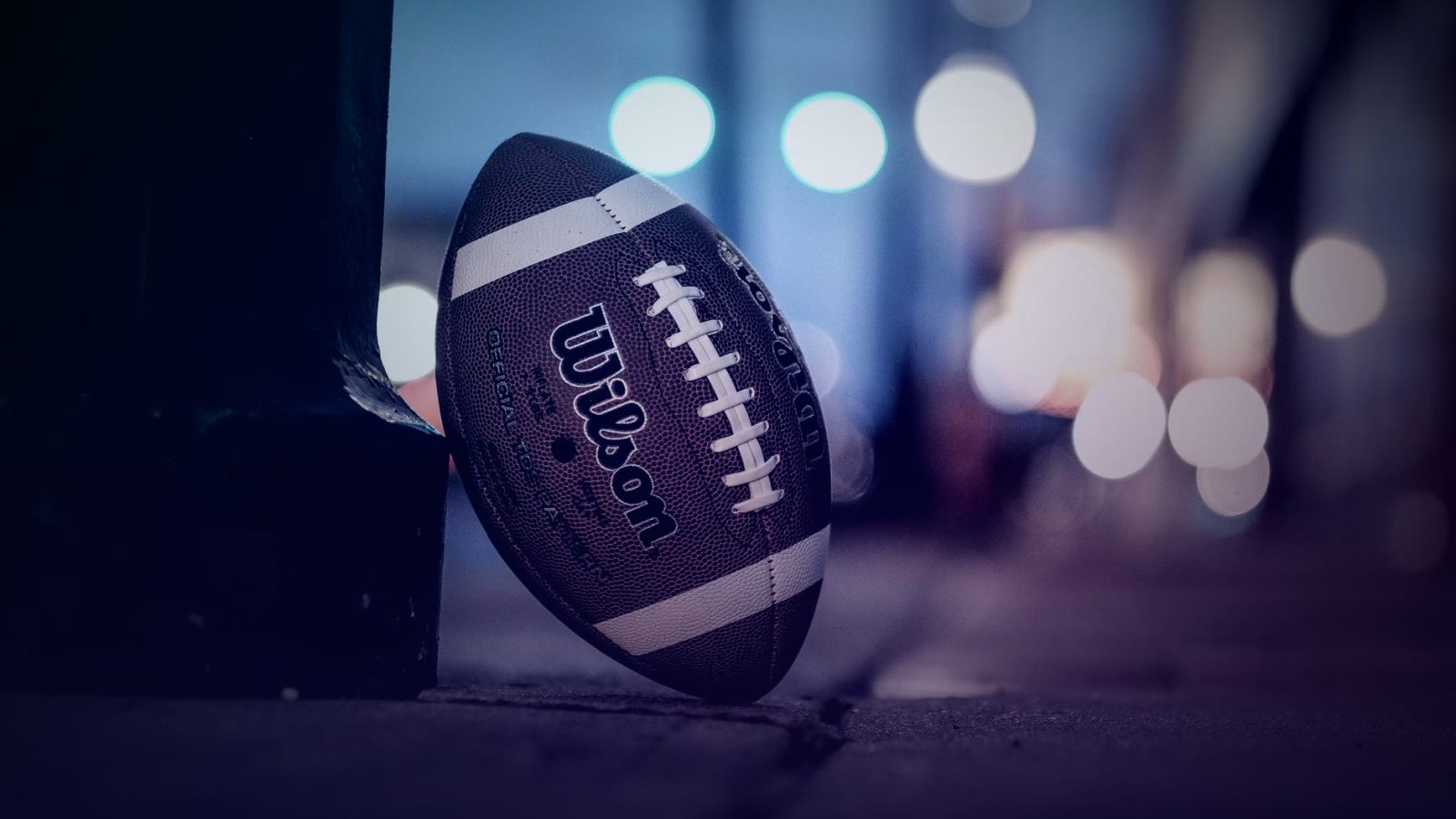 A Wilson American football rests on a city sidewalk at night with bokeh lights in the background.